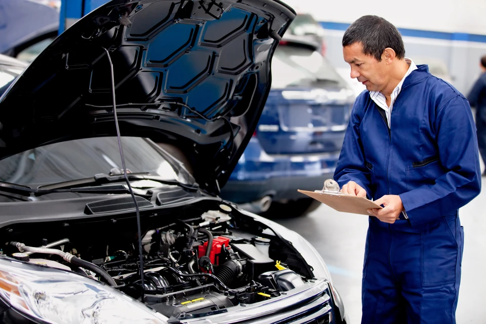 Mécanicien automobile en combinaison bleue examine un moteur, capot ouvert d'un véhicule noir dans un garage professionnel