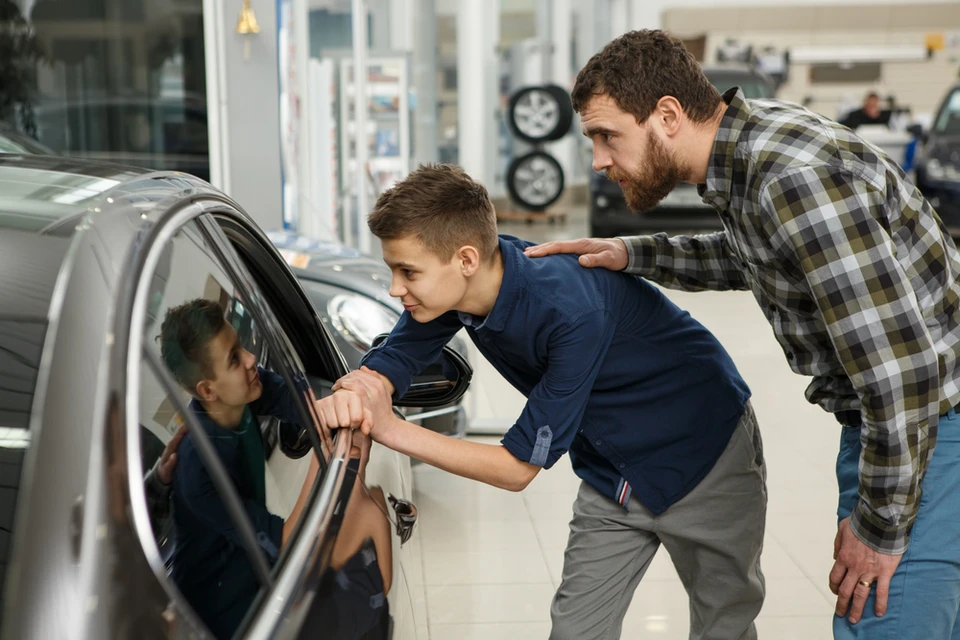 Un homme en chemise à carreaux supervise un jeune mécanicien en combinaison bleue qui explique les commandes d'un véhicule à un enfant assis au volant dans un garage automobile