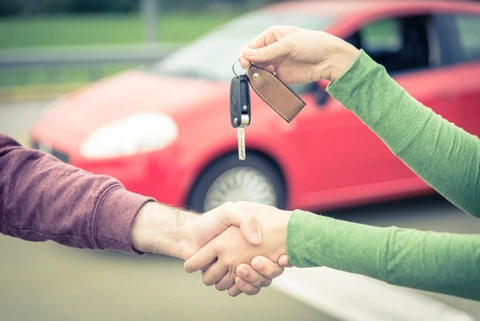 Scène extérieure de vente automobile : deux personnes effectuent une poignée de main lors de la remise des clés d'un véhicule rouge visible en arrière-plan sur un parking
