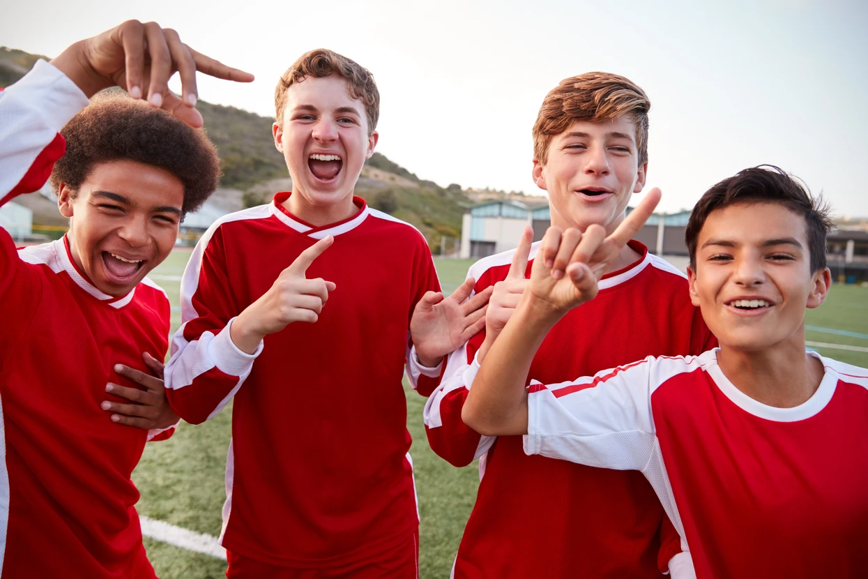 Quatre jeunes joueurs de football en maillots rouge et blanc célèbrent joyeusement sur un terrain de sport extérieur, faisant des gestes de victoire avec leurs mains levées et souriant