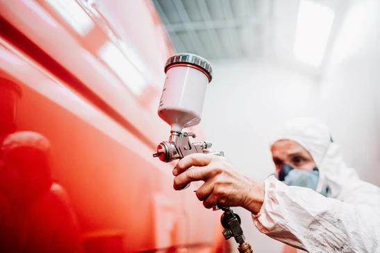 Atelier de carrosserie automobile intérieur avec ouvrier masqué appliquant peinture rouge sur carrosserie de véhicule