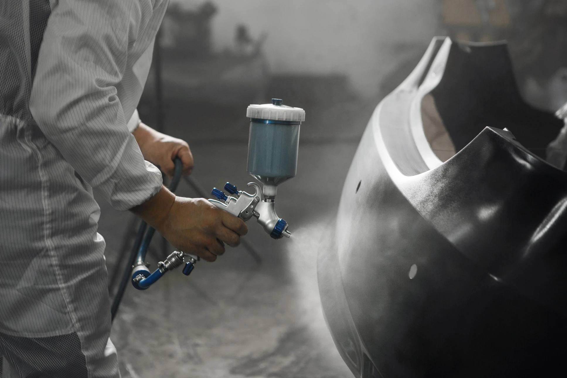 Atelier de carrosserie automobile où un technicien applique de la peinture bleue sur une pièce métallique à l'aide d'un pistolet à peinture pneumatique